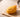 Golden crispy-coated food-shaped squishy with a curled strand coating and a small painted face, resting on a pale wooden table with soft warm daylight