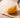 Golden crispy-coated food-shaped squishy with a curled strand coating and a small painted face, resting on a pale wooden table with soft warm daylight